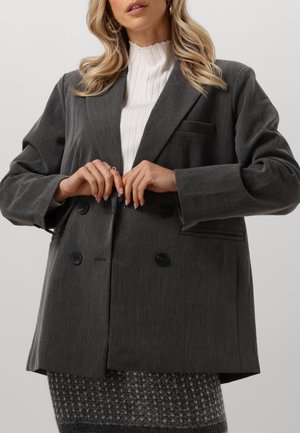 Grijze dubbelknop blazer van een gestructureerde stof, met grote zwarte knopen en een enkele borstzak. Gecombineerd met een witte geribbelde top.
