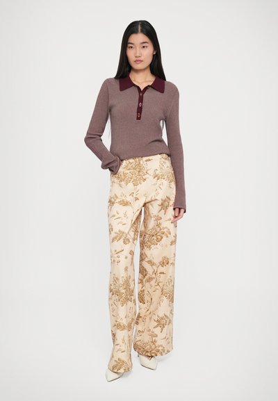 Smeđa rebrasta majica dugih rukava s bordo ovratnikom i gumbima, uparena s kremasto obojenim širokim hlačama s cvjetnim uzorkom.