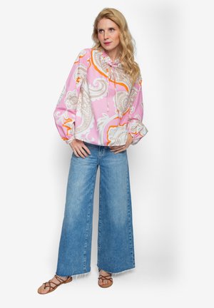 Hellrosa Bluse mit Paisley-Muster in Weiß und Orange, mit einem Kordelzug am Hals. Kombiniert mit weit geschnittenen blauen Jeans und braunen Sandalen.