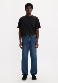 T-shirt noir à manches courtes, coupe décontractée, assorti à un jean bleu droit. Chaussures en daim noir. Design simple, sans motifs ni accents visibles.