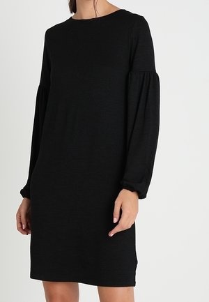 Femme portant une robe noire à manches longues avec des manches bouffantes froncées et un col rond sur un fond clair uni.