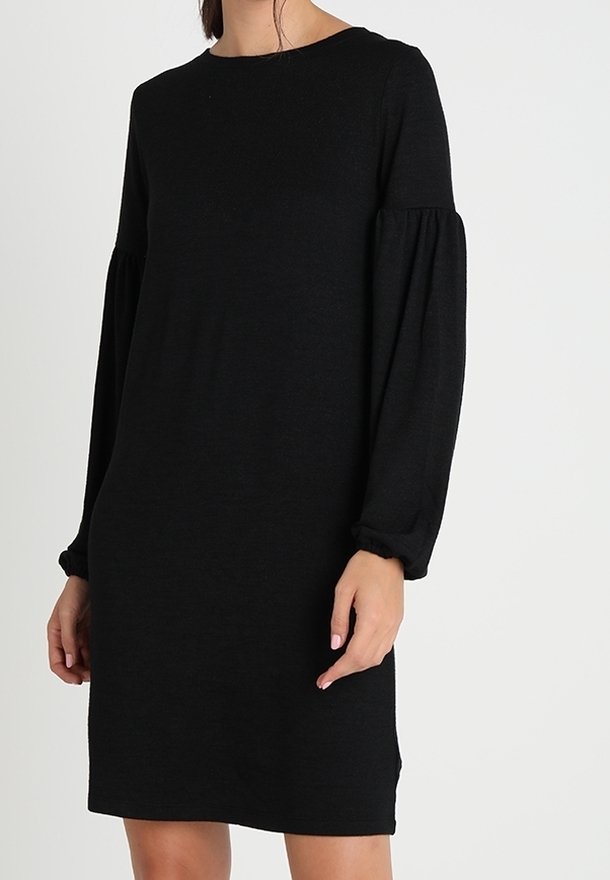 Femme portant une robe noire à manches longues avec des manches bouffantes froncées et un col rond sur un fond clair uni.