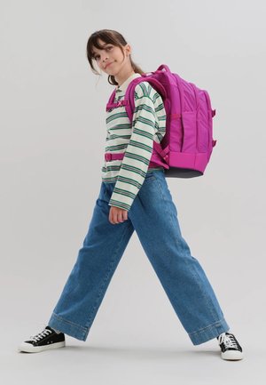 Young girl walking with a large bright pink backpack, wearing striped long-sleeve shirt, wide-leg jeans, and black sneakers.