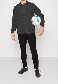 Pánská černá sportovní bunda s kulatým límcem, šedé tričko, černé kalhoty a černé fotbalové kopačky, držící vzorovaný fotbal.