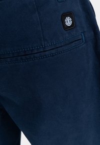 Marineblauwe broek met een achterzak voorzien van een zwart logo-embleem en contrasterende blauwe stiksels. De stof lijkt glad met een lichte glans.