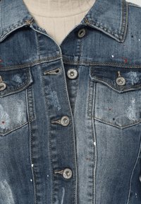 Chaqueta de mezclilla en un lavado médio con detalles desgastados, botones metálicos y acentos de pintura salpicada. Presenta bolsillos en el pecho y un diseño de cuello clásico.