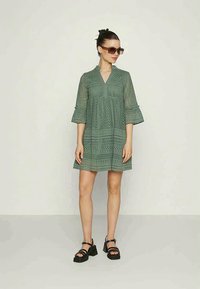 Žalia nėriniuota suknelė su išplatėjusiomis rankovėmis ir V formos iškirpte, dekoruota geometriniais raštais ir laisvu siluetu. Dėvima su juodomis platformomis.