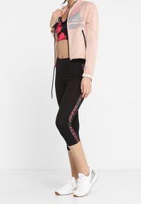 Giacca cropped rosa chiaro con tasca trasparente, reggiseno sportivo nero con stampa, leggings neri alla capri con striscia laterale rosa e sneakers bianche.