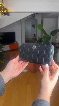 Zwarte gewatteerde leren portemonnee met zilveren accenten, voorzien van een ritssluiting en geometrisch patroon, vastgehouden door handen tegen een heldere interieurachtergrond.