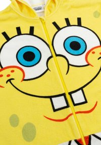 Tuta in pile gialla con il volto di un personaggio dei cartoni animati dotato di grandi occhi blu, denti quadrati e una bocca sorridente, con una chiusura centrale a zip.