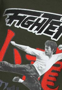 Olivengroen T-shirt met een zilveren "FIGHTER"-afbeelding en een zwart-witte afbeelding van een martial artist in een trappositie.