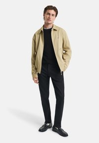 Braune Jacke mit Kragen, schwarzer Pullover darunter, schwarze Hose und schwarze Schuhe. Bemerkenswerte Nähte und Reißverschlussdetails an der Jacke.