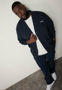 Cárdigan azul marino con botones y bolsillos sobre una camiseta blanca, combinado con pantalones cargo azules marinos y zapatillas blancas, contra un fondo neutro.