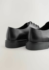 Zapatos de cuero negros con punta redondeada, superficie suave y diseño minimalista. Cuentan con una suela de goma negra baja y gruesa y cierre con cordones.
