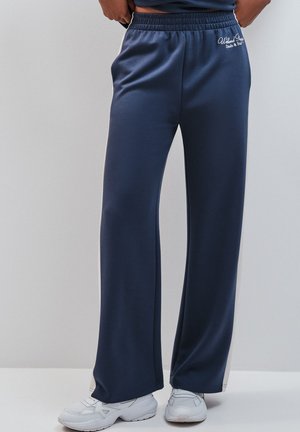 Personne portant un pantalon de jogging bleu marine ample avec des baskets blanches, debout devant un fond gris clair uni.