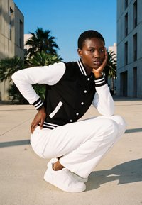 Mlada oseba s kratkimi lasmi, oblečena v črno-belo varsity jakno in bele hlače, ki čepi na prostem med stavbami z palmami.