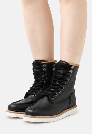 Bottines en cuir noir avec une texture lisse, semelle en caoutchouc blanc et lacets noirs. Dotées d'un col beige et d'œillets en métal.