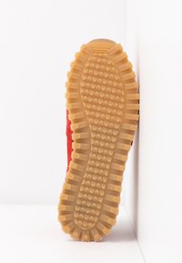 Punainen lenkkari, jossa on teksturoitu pinta ja kuminen ulkopohja aallokkomaisella kuviolla, joka näyttää kohotetut pidikkeet ja pyöristetyn kantapään.