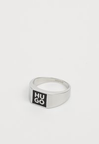 Anello in metallo argento con un logo nero rettangolare che presenta il testo "HUGO" in bianco. Superficie liscia, design piatto e bordi smussati.