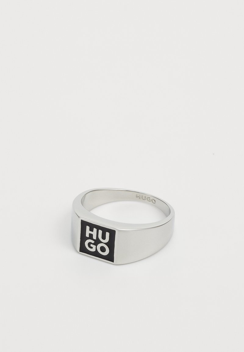 Anello in metallo argento con un logo nero rettangolare che presenta il testo "HUGO" in bianco. Superficie liscia, design piatto e bordi smussati.