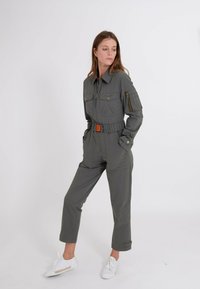 Grauer Utility-Overall mit einem taillierten Gürtel, Reißverschluss-Taschen und einem Kragen. Kombiniert mit weißen Turnschuhen und einer figurschmeichelnden Silhouette.