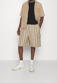 Krémová semišová košile s krátkým rukávem přes černé tričko, doplněná pruhovanými šortkami po kolena v béžové, modré a červené barvě. Bílé boty a ponožky.