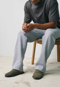 Szara koszulka z krótkim rękawem, jasnoszare dresowe spodnie i ciemnozielone klapki z prążkowaniem, noszone podczas siedzenia na drewnianym krześle. Widać wykładzinę.