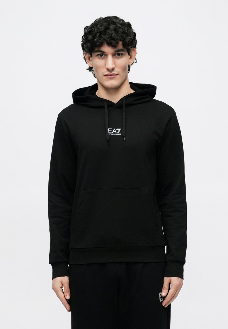 Joven con cabello rizado que lleva una sudadera con capucha negra de EA7 y pantalones negros a juego, de pie frente a un fondo blanco liso.