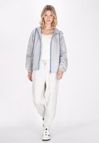 Giacca a vento grigio chiaro con zip e rifiniture blu, polsini elasticizzati e cappuccio, abbinata a pantaloni sportivi grigi e sneakers bianche.