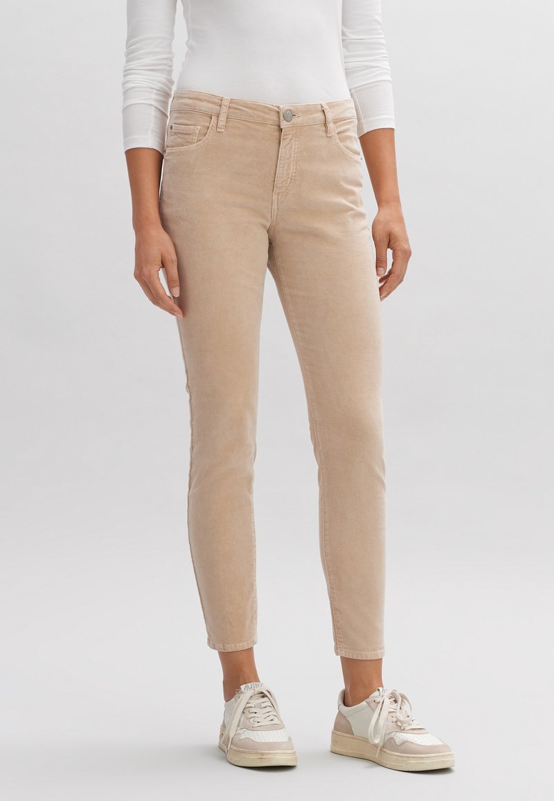 Pantaloni in corduroy beige con una vestibilità slim, dotati di tasche frontali, chiusura a bottone e un tessuto leggermente testurizzato. Abbinati a sneakers bianche.
