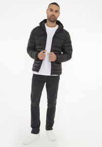 Chaqueta de plumas negra con capucha desmontable, diseño acolchado y cierre de cremallera, llevada sobre una camiseta blanca y combinada con jeans negros.