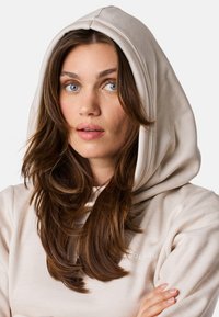 Felpa beige con un ampio cappuccio drappeggiato. Tessuto morbido e texturizzato. Presenta un marchio sottile sul petto. Maniche lunghe e larghe.