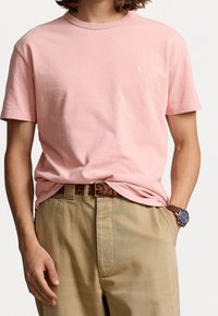 Kurzarm-T-Shirt mit Rundhalsausschnitt aus leichtem, rosa Baumwollstoff, das ein kleines gesticktes Logo auf der linken Brust trägt, kombiniert mit khakifarbene Hose und einem braunen Gürtel.