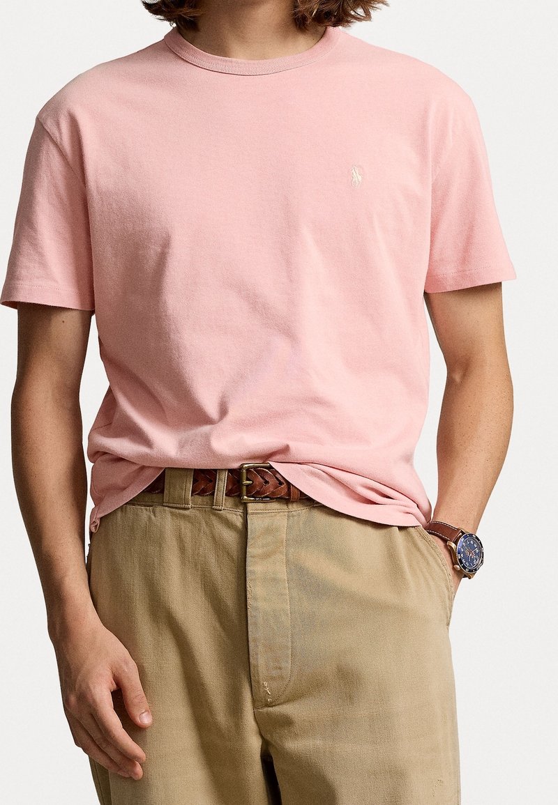 Kurzarm-T-Shirt mit Rundhalsausschnitt aus leichtem, rosa Baumwollstoff, das ein kleines gesticktes Logo auf der linken Brust trägt, kombiniert mit khakifarbene Hose und einem braunen Gürtel.