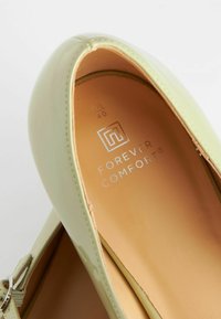 Grüne Lacklederschuhe mit abgerundetem Zehenbereich, die eine silberne Akzent-Schnalle und eine gepolsterte, tan-farbene Innensohle mit geprägtem Logo aufweisen.