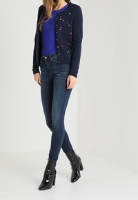 Navy knitted cardigan with colorful dots, worn over a blue top, paired with dark skinny jeans and glossy black heeled boots.