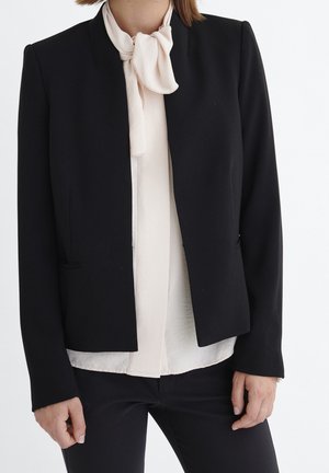 Blazer noir au design structuré, avec un col sans col, deux poches avant et une texture lisse, superposé à un chemisier clair.
