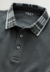 Grå polo t-shirt med en krave i ternet mønster, der har to grå knapper og et mærke, der står "NEXT." Glat tekstur og afslappet design.