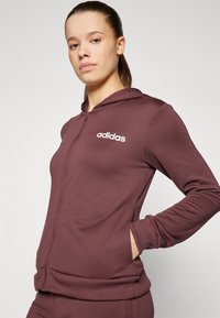 Maroon zip-up hoodie made of soft fabric, featuring a white Adidas logo on the left chest and ribbed cuffs and hem.