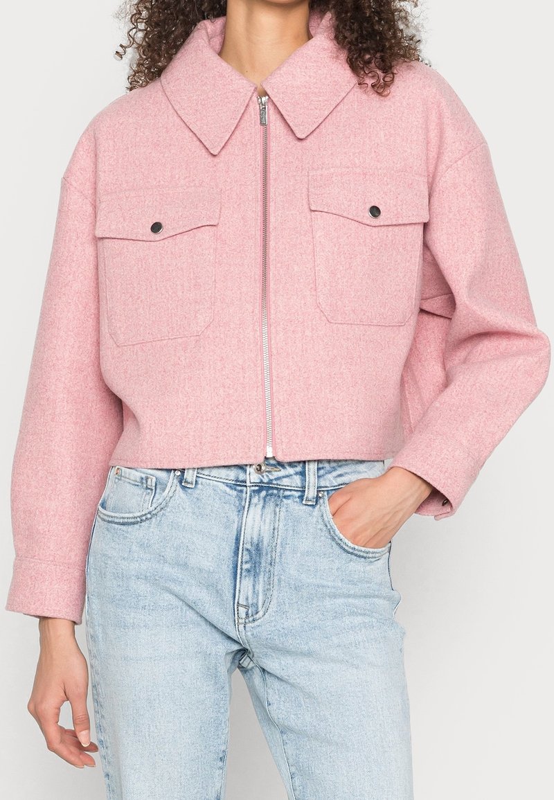 Femme portant une veste courte en laine rose avec fermeture éclair devant et poches à rabat, associée à un jean taille haute bleu clair.
