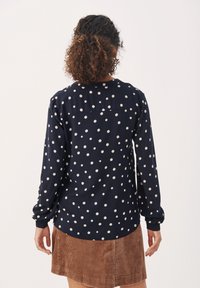 Femme aux cheveux bouclés portant une chemise à manches longues bleu marine à pois et une jupe en velours côtelé marron, debout dos à l'appareil photo.