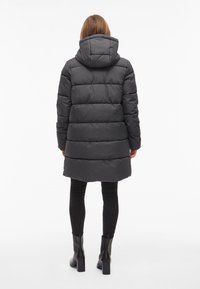 Puffer bunda v tmavo šedej farbe, s horizontálnym prešívaným dizajnom, kapucňou a dĺžkou po kolená. Kombinovaná s čiernymi legínami a členkovými čižmami.
