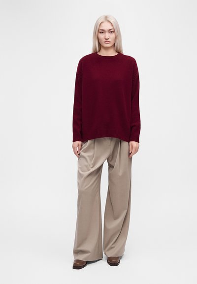 Personne debout aux longs cheveux blonds, portant un pull ample bordeaux, un pantalon beige large et des chaussures marron sur un fond uni.