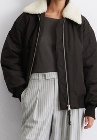 Svart bomberjacka med krämfärgad faux shearling-krage, quiltat mönster och silverfärgad dragkedja. Kombinerad med ljusgrå randiga byxor.