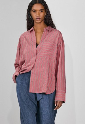 Žena s dlhými kučeravými vlasmi, oblečená v voľnej ružovej pruhovanej košeli so zapínaním na gombíky a modrých nohaviciach s vysokým pásom na jednoduchom sivom pozadí.