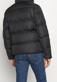 Zwarte pufferjack met een geborduurd ontwerp, met een hoge kraag en opvallende stiksels. Gecombineerd met blauwe denimjeans.