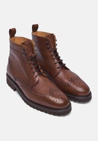 Stivali alla caviglia in pelle marrone, tomaia texturizzata, dettagli brogue, occhielli neri, cinque occhielli per i lacci e suola in gomma con battistrada.