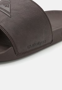 Sandalia deslizante de cuero marrón con una amplia correa superior que presenta un logo en relieve. Material texturizado y bordes con costuras.