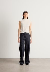 Krém színű ujjatlan felső, magas nyakú; laza szabású sötét farmernadrág; sima textúra; fekete öv; minimalista kialakítás minták nélkül.