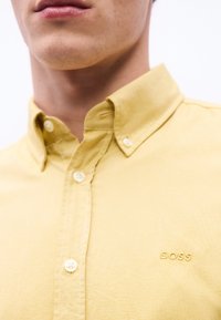 Κοντινό πλάνο ενός ατόμου που φοράει πουκάμισο κίτρινο με κουμπιά και γι ακά, με την επιγραφή "BOSS" κεντημένη στο στήθος.